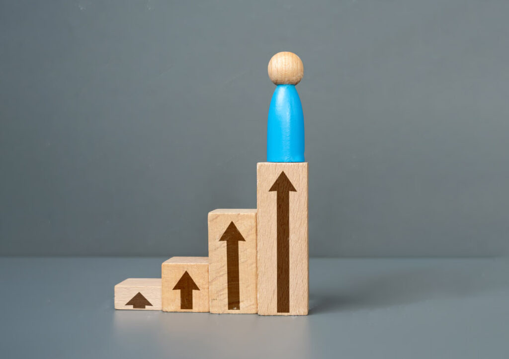 木製の積み木が階段状に配置され、各ブロックには上向きの矢印が描かれている。最上段のブロックには青の駒が立っている。背景はグレー。